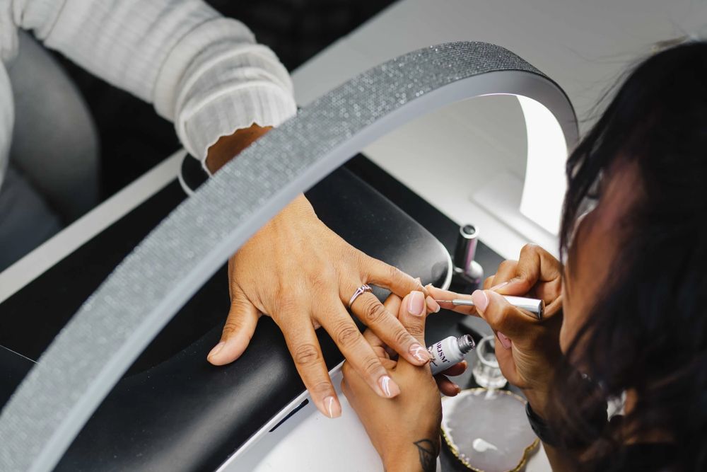 Thiara brushing a thin layer of gel nail polish onto a client's natural nails under the lamp during a gel manicure at TLC Nails and Esthetics in Nottingham, MD