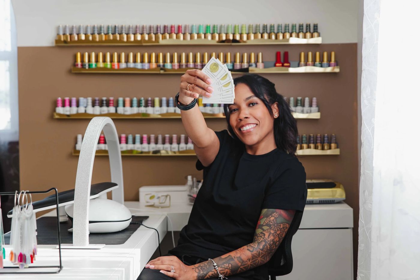 Thiara holding up Gel X service cards at her manicure desk, LED lamp and racks of gel polish behind her, TLC Nails and Esthetics in Nottingham, MD