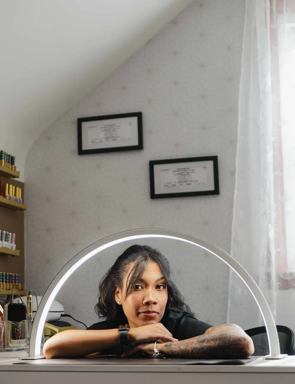 Thiara, the licensed esthetician at TLC Nails and Esthetics in Nottingham, MD, resting her folded arms on her white desk inside a half-moon lamp, with her two framed Maryland licenses on the patterned wall behind her