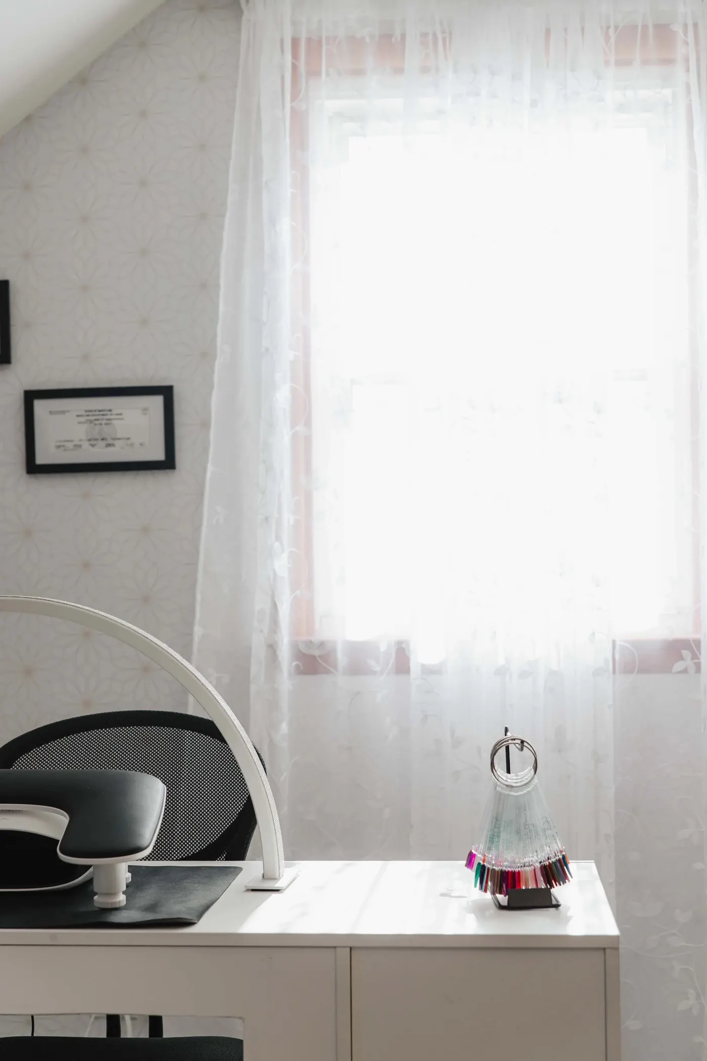 The empty private suite at TLC Nails and Esthetics in Nottingham, MD in daylight: one white work desk, a ring lamp, a gel color swatch fan, and Thiara's framed Maryland license on the wall beside a sheer curtained window