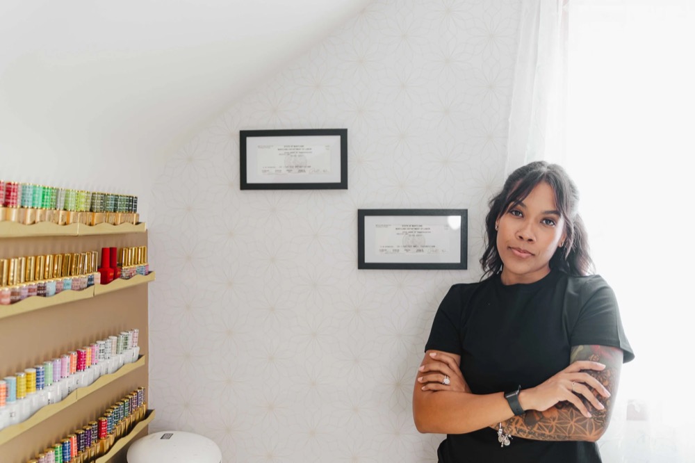 Thiara, the licensed esthetician and nail technician at TLC Nails and Esthetics in Nottingham, MD, standing beside her two framed Maryland licenses and her wall of gel polish