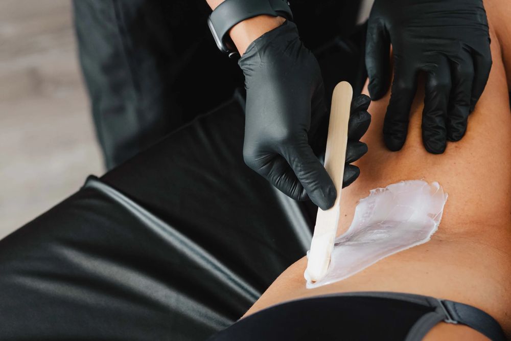 Gloved hands spreading warm soft wax with a wooden spatula along a client's bikini line at TLC Nails and Esthetics in Nottingham, MD