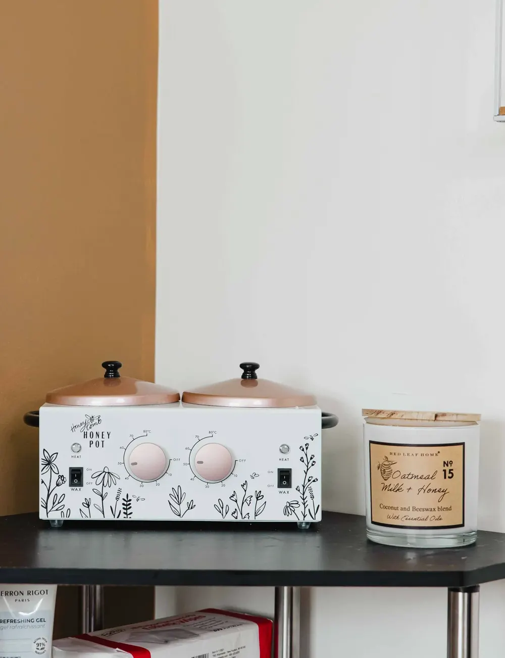 A two-pot Honey Pot wax warmer with rose gold lids beside an Oatmeal Milk and Honey candle on the black shelf of the waxing station at TLC Nails and Esthetics in Nottingham, MD